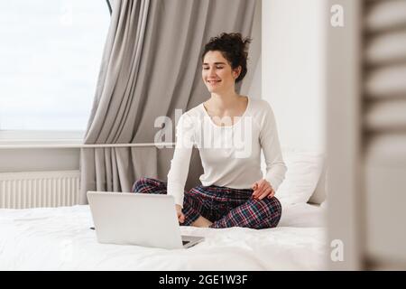 Die lächelnde Frau, die mit gekreuzten Beinen vor einem Laptop auf dem Bett sitzt, während sie in einem hellen Raum auf seine Kamera blickt Stockfoto