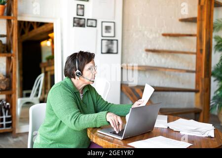 Eine ältere Frau arbeitet zu Hause vor einem Laptop-Monitor mit Papieren. Heimbüro. Sie geht durch Steuererklärungen und prüft Rechnungen. Stockfoto