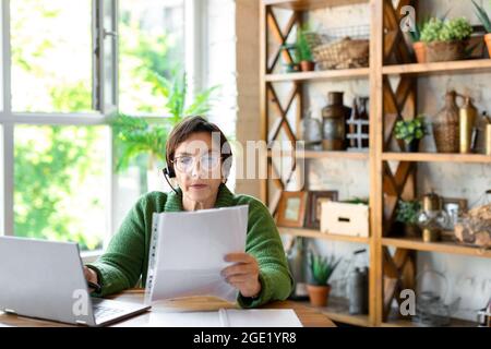 Eine ältere Frau arbeitet zu Hause vor einem Laptop-Monitor mit Papieren. Heimbüro. Sie geht durch Steuererklärungen und prüft Rechnungen. Stockfoto