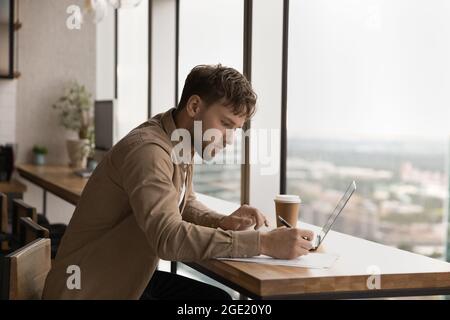Serious Mann sitzen am Arbeitsplatz Verwenden Sie Laptop notiert in Papier Stockfoto