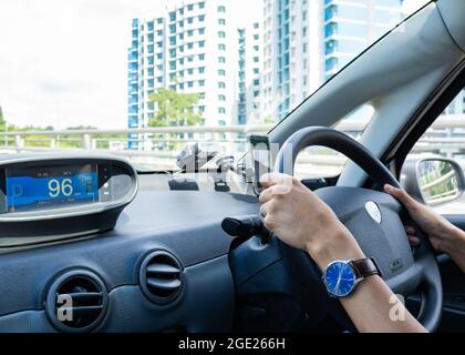 Armaturenbrett eines Elektroautos, das von einem jungen Mann mit brauner Haut gefahren wird (nur Hände sind sichtbar).Batterieanzeige des Autos ist sichtbar. Singapur Stockfoto