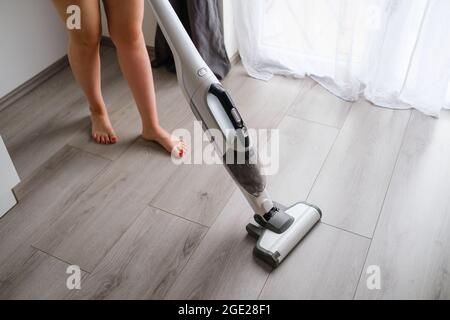 Frau, die einen Boden mit kabellosem Staubsauger im Zimmer mit moderner Einrichtung reinigt. Einfache Entfernung von Schmutz. Stockfoto