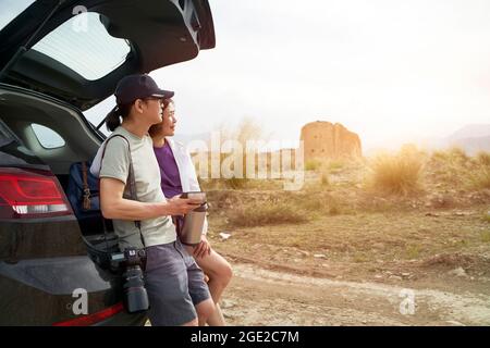 das asiatische Paar lehnte sich an der Rückseite des Autos und schaute auf die Aussicht, während es Kaffee an einem verlassenen historischen Ort trank Stockfoto