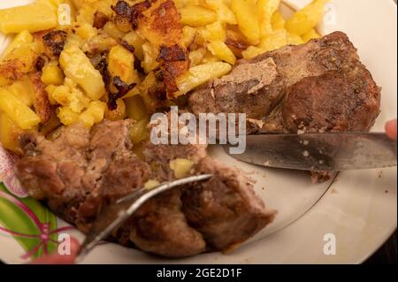 Jemand schneidet mit einem Messer und einer Gabel ein Stück saftiges Braten auf einem Teller gebratener Kartoffeln. Nahaufnahme Selektiver Fokus Stockfoto