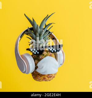 Kreative lustige Ananas Gesicht trägt Brille Kopfhörer und schützende medizinische Maske. Ruhende schwebender Ananas Gesicht auf Farbe gelb Sommer Stockfoto