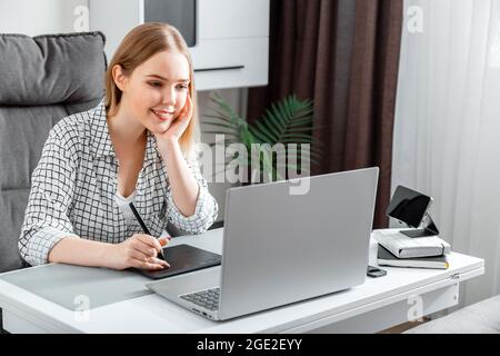 Lächelnde, fröhliche Designerin, die im Home Office mit einem GrafikTablet und einem Laptop arbeitet. Freiberufliche Retuscherin Illustratorin junge kaukasische blonde Frau Stockfoto