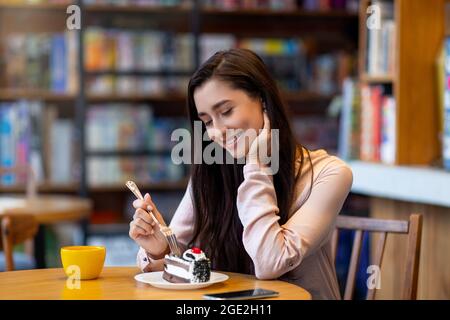 Nachtisch. Fröhliche arabische Frau, die Dessert genießt und Kaffee trinkt, während sie im Café sitzt, freier Platz Stockfoto