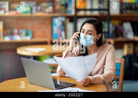 Vielbeschäftigte arabische Freiberuflerin in Gesichtsmaske, die am Handy sprach und Papiere überprüfte, am Laptop im Café arbeitete Stockfoto