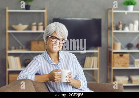 Porträt einer glücklichen, reifen Frau, die auf der Couch sitzt, eine gute Tasse Kaffee genießt und lächelt Stockfoto