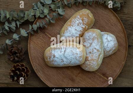 Vier hausgemachte süße, cremefarbene, weiche Brötchen und bestreut mit Puderzucker auf einem runden Holzteller über einem rustikalen Holztisch. Bäckerei- und Dessertkonzept, Selecti Stockfoto