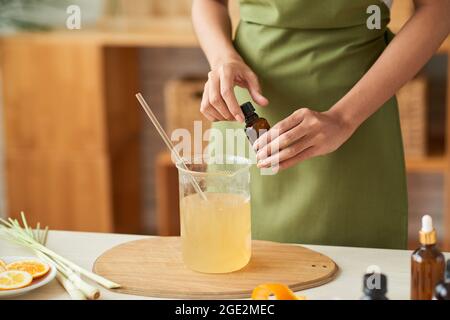 Hände einer Frau, die ätherisches Öl in ein Glas geschmolzener Seifenbasis eingibt, wenn sie zu Hause Reinigungsstäbe macht Stockfoto