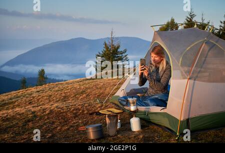 Junge Wandererin fotografiert mit dem Smartphone bei Sonnenaufgang in den Bergen von ihrem Zelt aus neben Kochgeräten während des Wanderns. Im Hintergrund Bäume und Nebel am Fuße der Berge. Stockfoto
