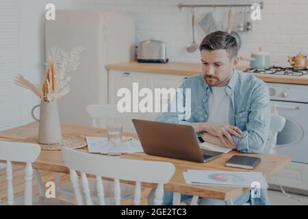 Fokussierter junger Freiberufler, der am Küchentisch sitzt und am Laptop arbeitet Stockfoto