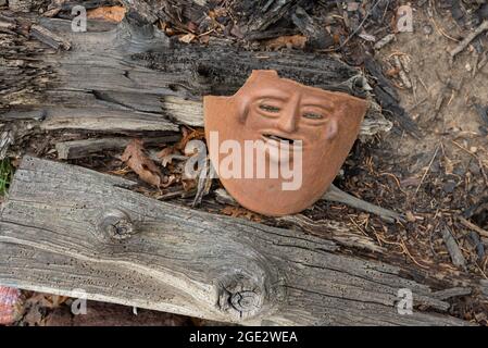 Eine weggeworfene zerbrochene Tonmaske, die auf dem Boden auf einem verrottenden Baumstamm, getrockneten Blättern und Zweigen liegt. Stockfoto