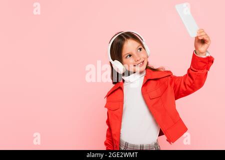 Fröhliches und trendiges Mädchen, das Selfie in Kopfhörern auf rosa isoliert macht Stockfoto