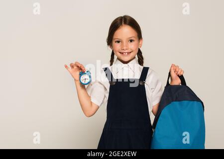 Lächelndes Schulmädchen hält blauen Rucksack und kleinen Wecker isoliert auf grau Stockfoto