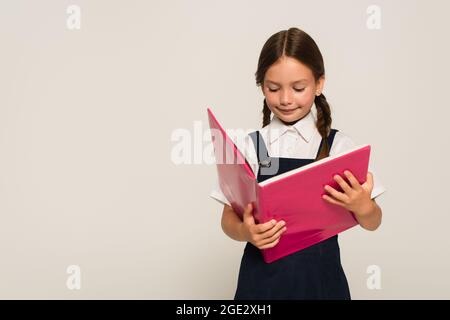 Lächelndes Mädchen in der Schuluniform liest Notizbuch isoliert auf grau Stockfoto