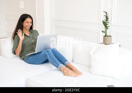 Glücklich lächelnde indische Frau mit einem Laptop hat Videoanruf, virtuelle Sitzung. Eine junge Mischrennerin, die sich auf der weißen bequemen Couch niederlegt und fröhlich in die Webcam winkt Stockfoto