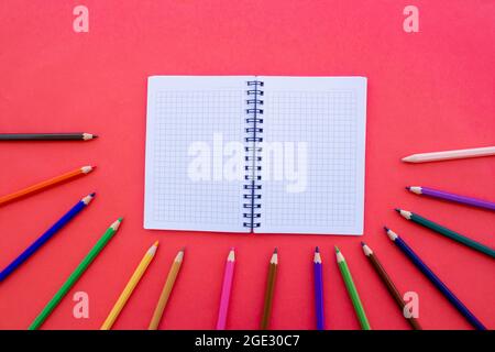 Bunte Buntstifte und leerer Notizblock auf rotem Hintergrund isoliert. Zurück zum Schulkonzept. Draufsicht, Kopierbereich, flach liegend. Stockfoto