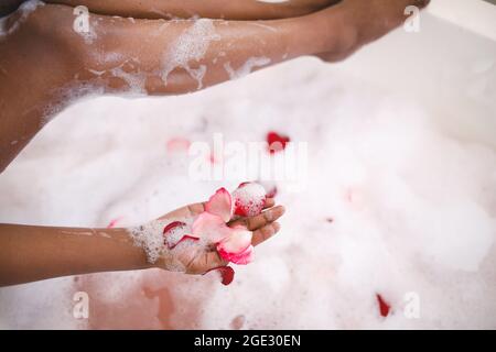 Nahaufnahme einer afroamerikanischen Frau, die sich in einem Schaumbad verwöhnen lässt und Rosenblätter hinzufügt Stockfoto