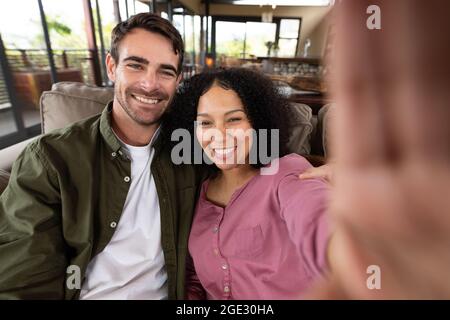 Porträt eines glücklichen, vielfältigen Paares, das im Wohnzimmer auf dem Sofa sitzt und Selfie macht und lächelt Stockfoto