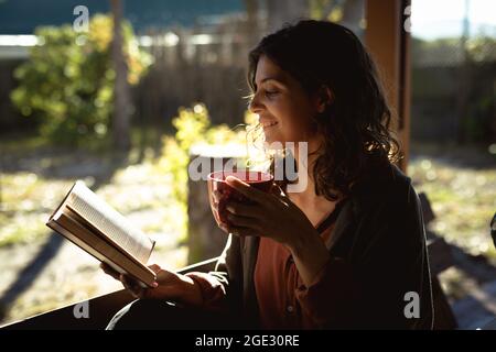 Frau mit gemischter Rasse liest Buch und trinkt Kaffee im sonnigen Garten Stockfoto