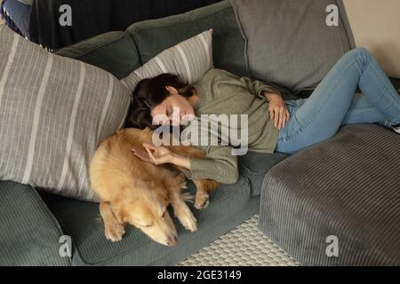 Lächelnde kaukasische Frau im Wohnzimmer, die mit ihrem Hund auf dem Sofa liegt Stockfoto
