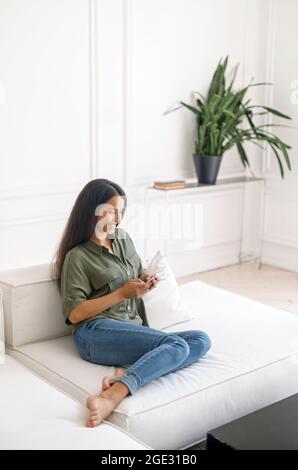 Die vertikale Aufnahme einer lächelnden jungen indischen Frau unterhält sich gerne auf dem Smartphone, das zu Hause auf der Couch sitzt. Dunkelhaarige, fröhliche Mischrasse-Frau hält Handy und SMS online Stockfoto