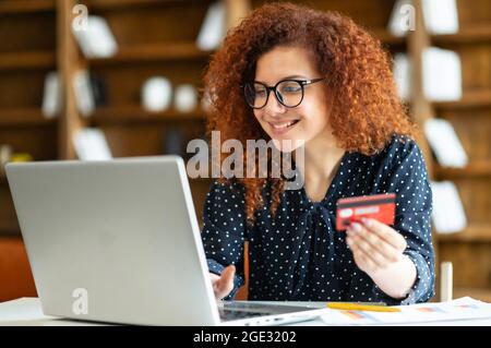 Lächelnde Geschäftsfrau mit lockiger Frisur hält Kreditkarte, um Online-Transaktionen zu tätigen, schaut auf Laptop, kauft gewünschte Artikel, macht Kauf online, Einkaufen von zu Hause aus Konzept Stockfoto