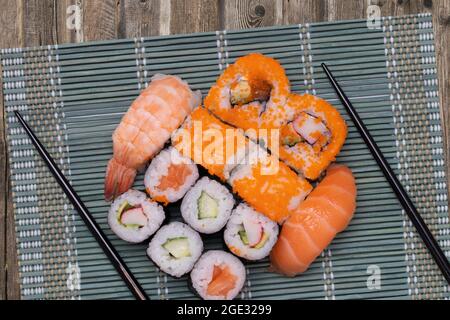 Nahaufnahme verschiedener Sushi-Rollen mit Lachs, Sashimi und anderen Scheiben rohen Fischs und einem Paar hölzernen Essstäbchen, die auf einer grünen Bambusmatte serviert werden Stockfoto