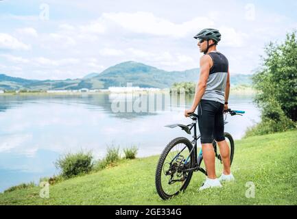 Ein Radfahrer mittleren Alters, der in Radbekleidung, Helm und Sonnenbrille gekleidet ist, mit dem Fahrrad auf einem Grashügel, der die See- und Berglandschaft genießt. Out-of Stockfoto