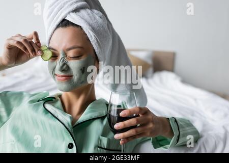 Fröhliche sianische Frau in Tonmaske, die Gurkenscheiben auf das Auge aufführt, während sie ein Glas Rotwein hält Stockfoto