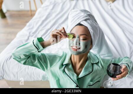 Fröhliche asiatische Frau in Tonmaske, die Gurkenscheiben auf das Auge aufgibt, während sie ein Glas Rotwein hält Stockfoto