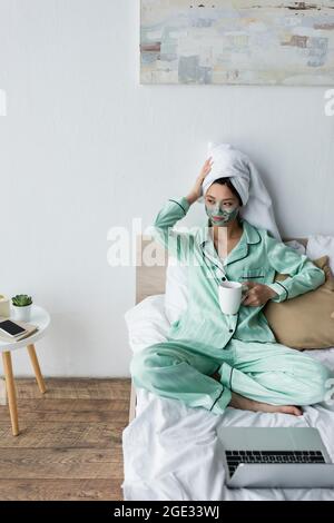 asiatische Frau in Lehmmaske und Schlafanzug berühren Handtuch auf dem Kopf, während sie in der Nähe Laptop auf dem Bett sitzen Stockfoto