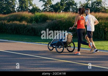 Die junge Familie läuft mit dem Baby-Joggingwagen in Bewegung und genießt das Training und den Sommertag im Stadtpark. Rückansicht Stockfoto