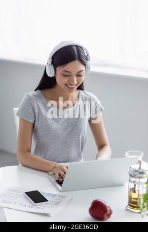 Lächelnde Asiatin mit Kopfhörern, die auf dem Laptop neben der Teekannen und dem Smartphone auf dem Schreibtisch tippt Stockfoto