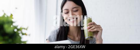 Hübsche asiatische Frau lächelt, während sie ein Glas leckeren Smoothie, Banner hält Stockfoto