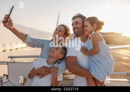 Fröhliche Touristen, schöne Familie, die Selfie macht, während sie im Freien zusammen steht, um bei Sonnenuntergang in das Flugzeug zu steigen Stockfoto