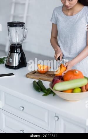 Teilansicht einer asiatischen Frau, die Kürbis in der Nähe von Gemüse, Shaker und Smartphone mit leerem Bildschirm schneidet Stockfoto