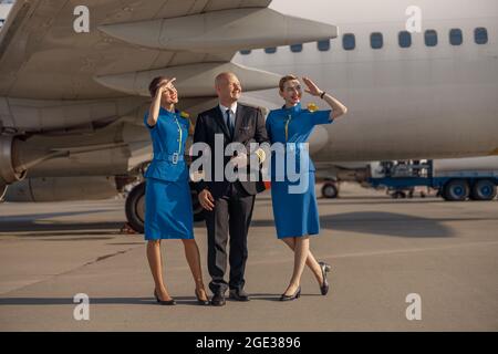 In voller Länge ein glücklicher Pilot, der an einem sonnigen Tag zusammen mit zwei Stewardessen in hellblauer Uniform vor einem Flugzeug steht Stockfoto