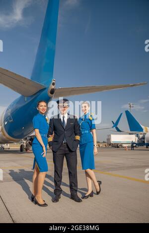 In voller Länge ein fröhlicher Pilot und zwei hübsche Stewardessen, die vor einem Flugzeug stehen und die Kamera anlächeln Stockfoto