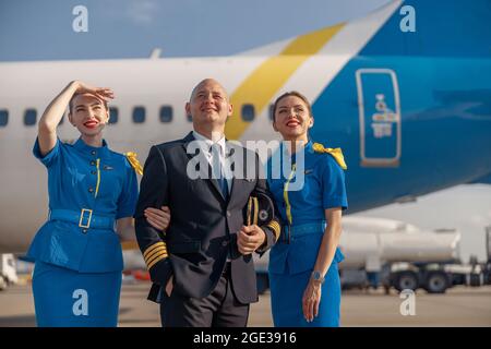Glücklicher Pilot und zwei hübsche Stewardessen stehen vor einem Flugzeug und lächeln nach der Landung weg Stockfoto