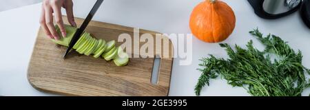 Teilansicht einer Frau, die frische Zucchini auf einem Schneidebrett in der Nähe eines reifen Kürbisses auf dem Tisch schneidet, Banner Stockfoto