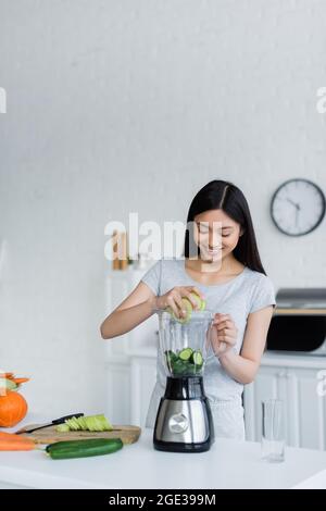 Glückliche asiatische Frau, die in Scheiben geschnittene Gurken in den Shaker neben frischem Gemüse und Schneidebrett einfügte Stockfoto