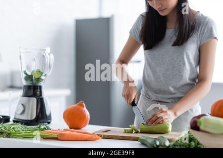 Teilansicht einer Frau, die Zucchini in der Nähe des Mixers, rohen Kürbis und Karotten auf dem Tisch schneidet Stockfoto