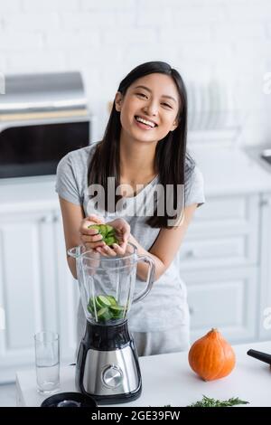 Fröhliche asiatische Frau, die in Scheiben geschnittene Gurken in den Mixer gab, während sie das Frühstück in der Küche zubereitete Stockfoto