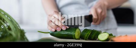 Beschnittene Ansicht einer verschwommenen Frau, die frische Gurken auf einem Schneidebrett, einem Banner, schneidet Stockfoto