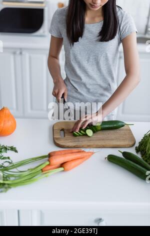 Teilansicht einer Frau, die in der Küche Gurken in der Nähe von frischen Karotten und Kürbis schneidet Stockfoto