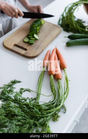 Teilansicht einer Frau, die frische Gurken in der Nähe von rohen Karotten auf dem Tisch schneidet Stockfoto