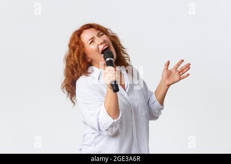 Emotionen, Lifestyle und Freizeitkonzept. Leidenschaftliche und sorglose Rothaarige Performerin, Frau mittleren Alters, die Lied im Mikrofon singt, Sängerin Stockfoto